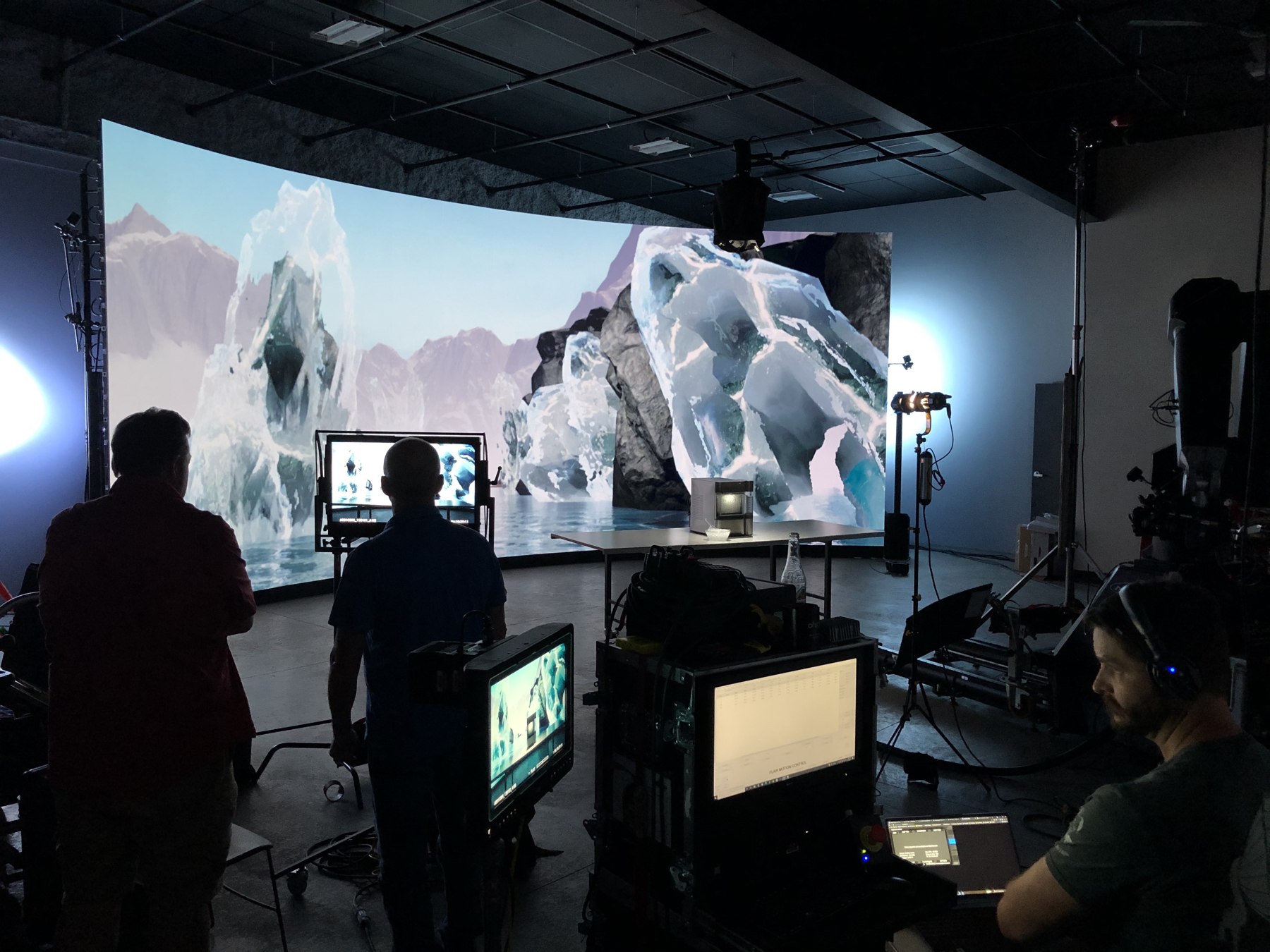 Wide studio view with crew, cameras and practical objects in front of a curved LED wall displaying mountains